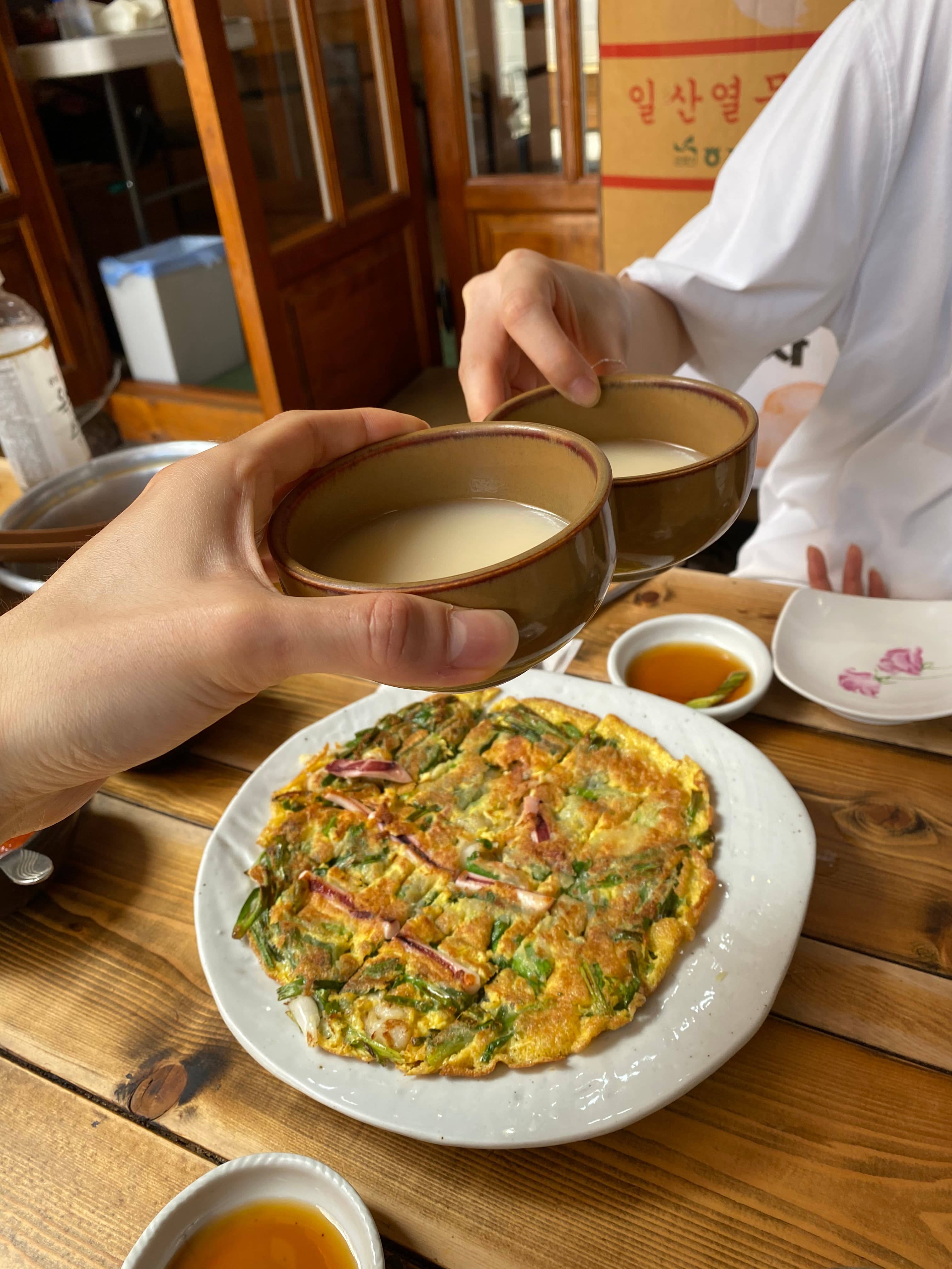 Friends raising bowls of makgeolli over a freshly cooked Korean pajeon