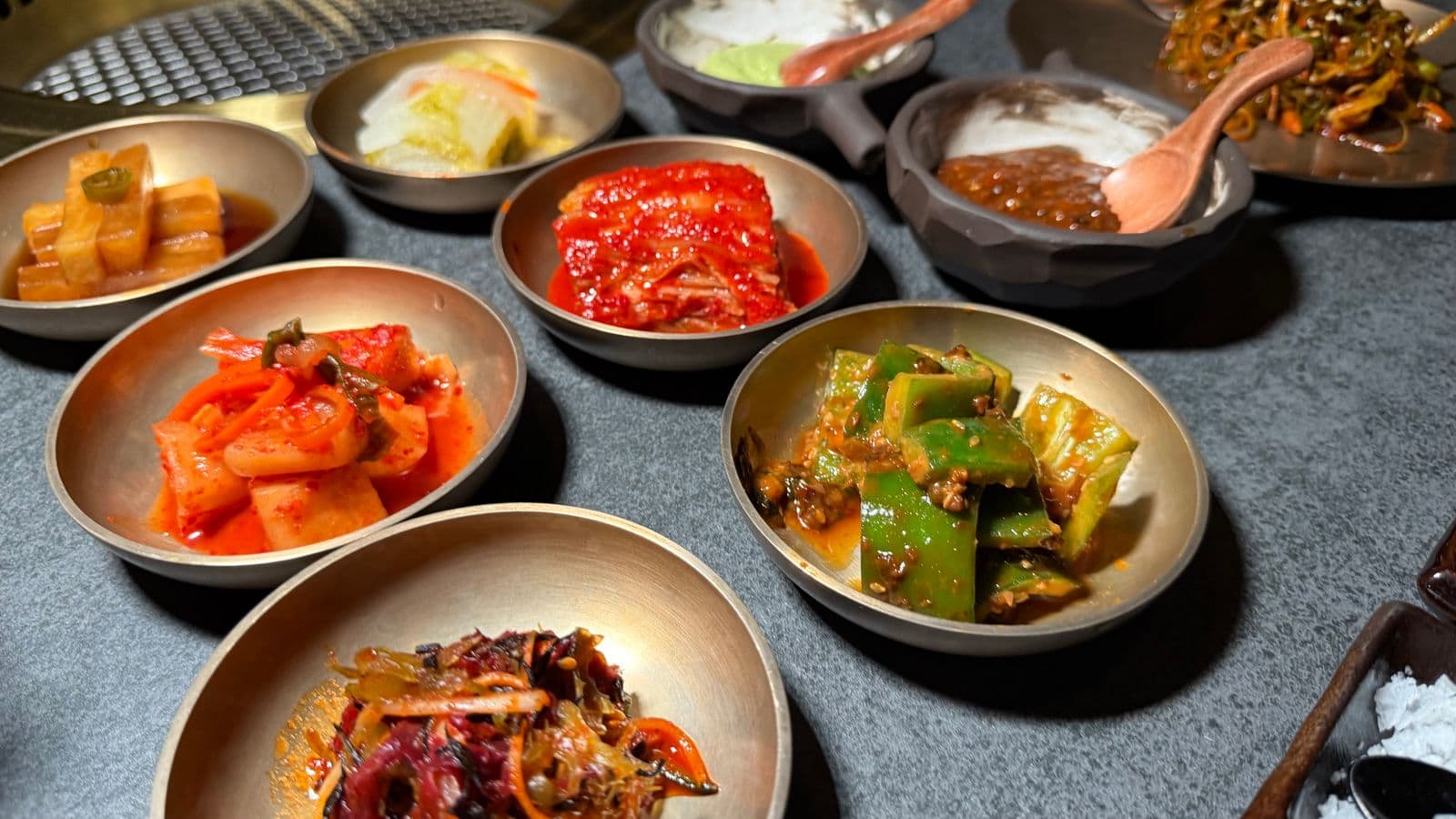 Korean banchan side dishes in metal bowls on a table — kimchi, seasoned vegetables, and ssamjang beside a grill