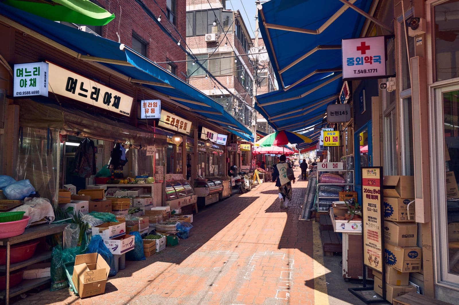 A Seoul traditional market alley under blue awnings, with Korean shop signs and vegetables stacked in crates
