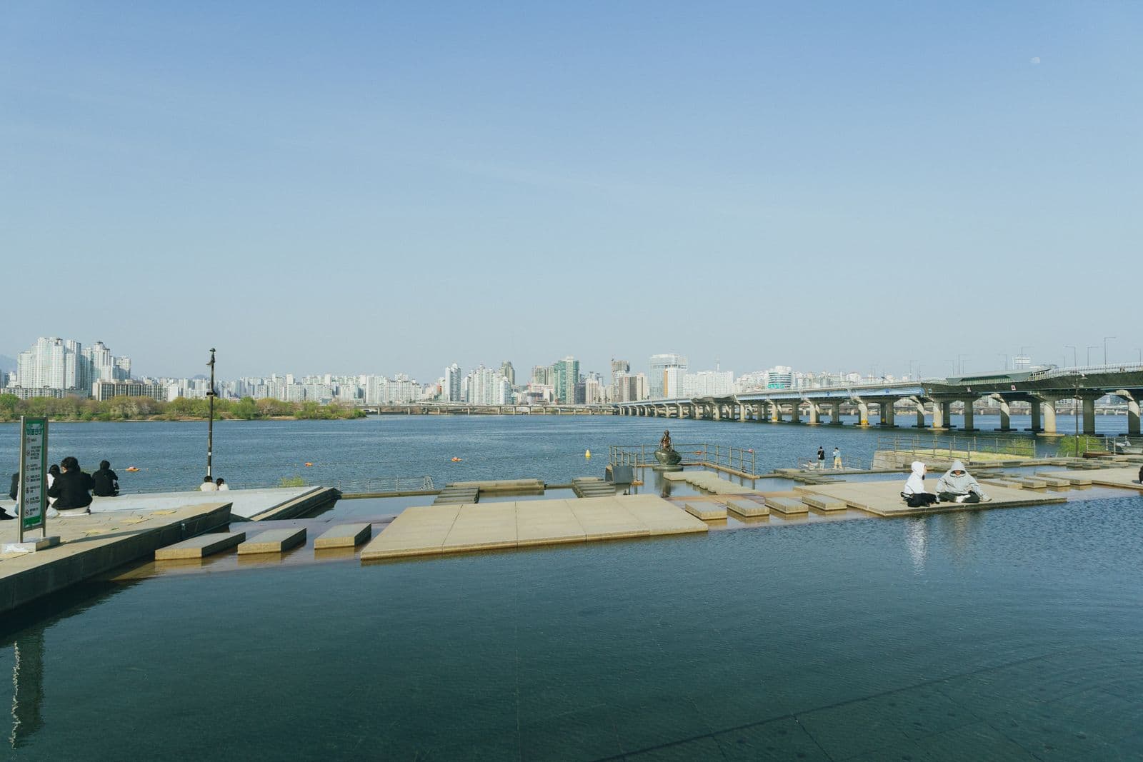 People sitting on riverside steps beside the Han River with Seoul and a bridge behind them
